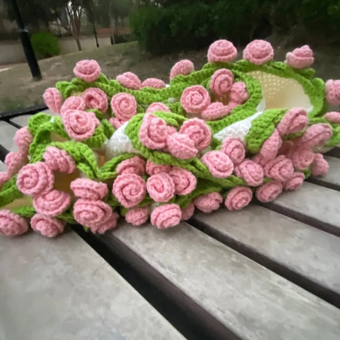 A handmade crochet blanket with soft pink 3D rose flowers and green leafy vines, draped over a wooden park bench in a serene outdoor setting