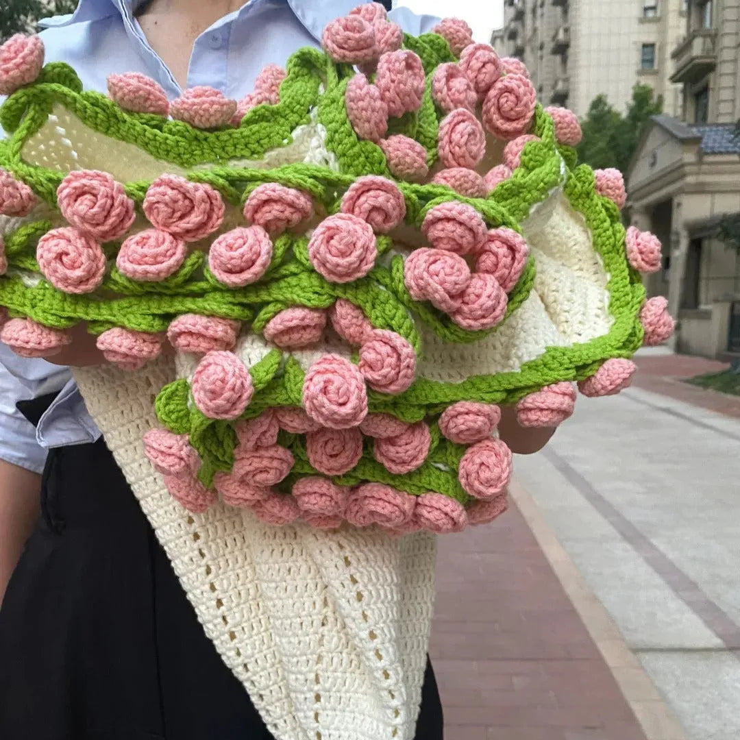A handmade bouquet-style crochet blanket with soft pink rose flowers and green leafy vines, wrapped in a cream cone base and held by a person on a city sidewalk