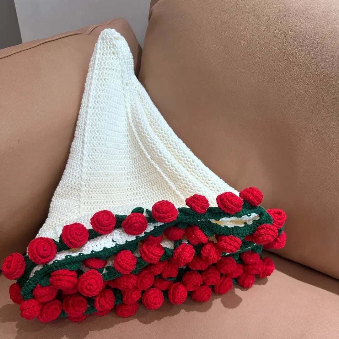 A handmade white crochet blanket folded into a bouquet shape, featuring dense red crochet roses and green leaves at the base, resting on a beige sofa for a cozy presentation