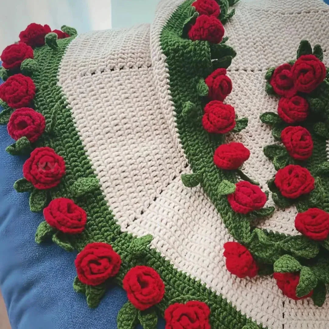 A handmade crochet blanket featuring red rose flowers and green leaves along the edges, laid out flat to showcase the round design and intricate stitching details