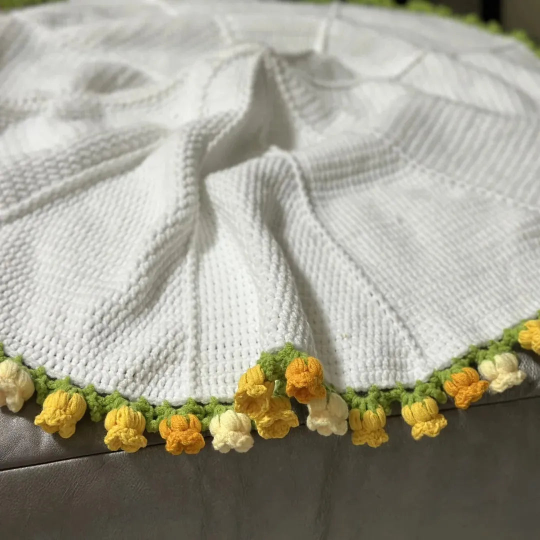 A round handmade white crochet blanket with delicate yellow and cream tulip-style 3D flower trim and green leafy edge, draped naturally on a soft gray surface