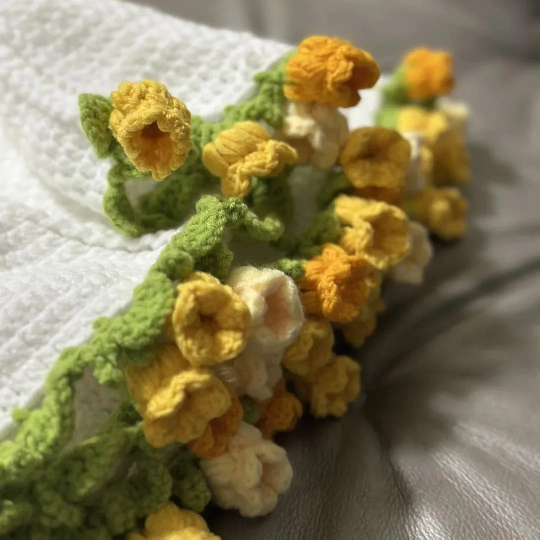 Macro shot of yellow and cream 3D crochet flowers with green leafy edges on a white handmade blanket, displayed on a soft gray leather sofa