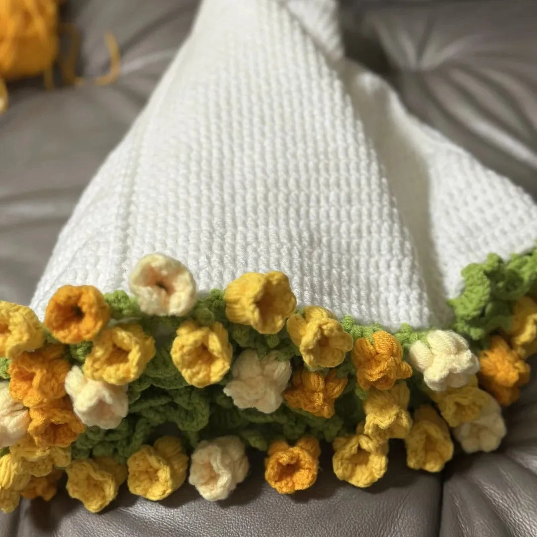 Detail view of a handmade white crochet blanket featuring delicate yellow and cream 3D flower edging with green leaves, resting on a gray tufted sofa