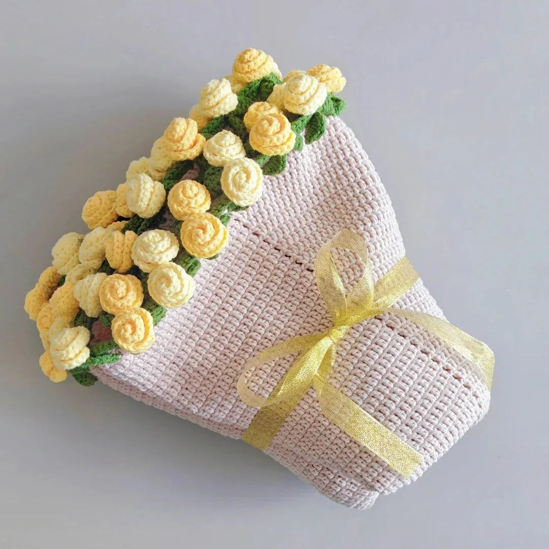 A handmade crochet bouquet blanket styled with soft yellow rose flowers and green leaves, wrapped in a cream cone with a golden ribbon bow, displayed on a light gray background